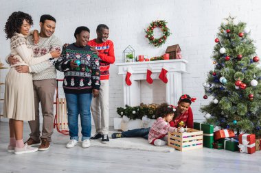 Mutlu Afro-Amerikan ailesi oturma odasındaki Noel ağacının yanında oynayan erkek ve kız kardeşlerine bakıyor.