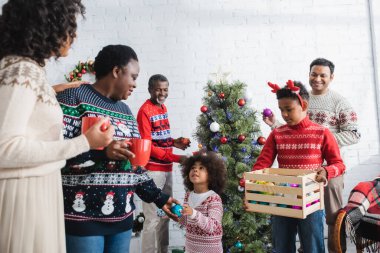 Ren geyiği boynuzları takan Afrikalı Amerikalı çocuk aile ve noel ağacının yanında süslü takılar taşıyan ahşap bir kutu tutuyor.