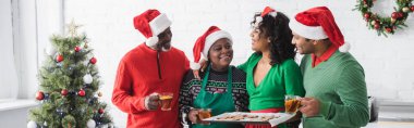 Orta yaşlı Afrikalı Amerikalı bir kadın elinde Noel kurabiyeleriyle birlikte mutlu bir aile ve Noel ağacının yanında pankart tutuyor.