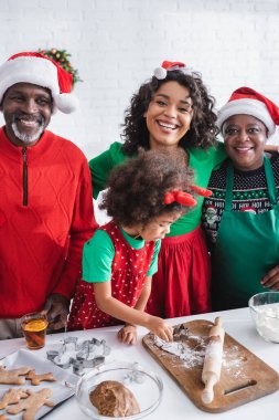 Ren geyiği boynuzlu Afrikalı Amerikalı kız Noel Baba şapkalı mutlu ailenin yanında noel kurabiyeleri hazırlıyor.