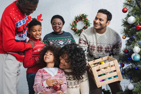 Gülümseyen adam elinde mücevher dolu tahta kutuyla Noel ağacının yanında ve mutlu Afro-Amerikan ailesi