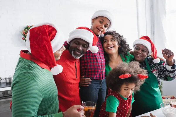 Noel Baba şapkalı neşeli Afro-Amerikan ailesi mutfakta kucaklaşıp gülümsüyor.