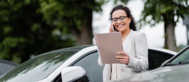 Kağıt klasörlü gülümseyen Afrikalı Amerikalı menajer araba parkında akıllı telefondan konuşuyor.