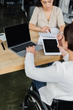 Tekerlekli sandalyedeki iş kadını, Afrikalı Amerikalı meslektaşının yanında dijital tablet ve dizüstü bilgisayarla çalışıyor.