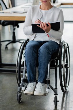 Tekerlekli sandalyedeki iş kadınının ofiste çalışırken deftere yazdığı kısmi görünüm