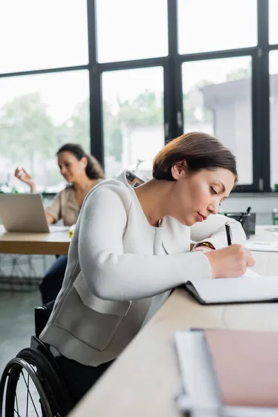 Mujer Negocios Con Discapacidad Escribiendo Cuaderno Cerca Borrosa Colega Afroamericano —  Fotos de Stock