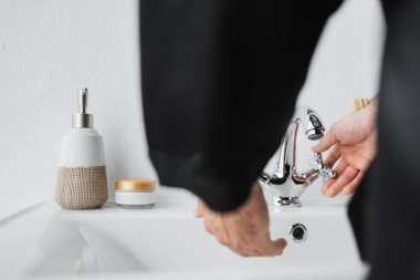 Banyonun lavabosunun yanındaki muslukta musluk açan adamın görüntüsü. 
