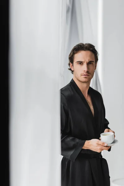 Young man in silk robe holding coffee near curtains at home