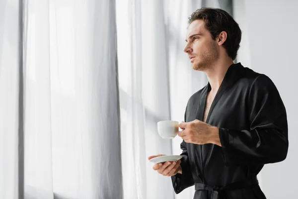 Young man in black robe holding coffee cup and saucer near curtains at home