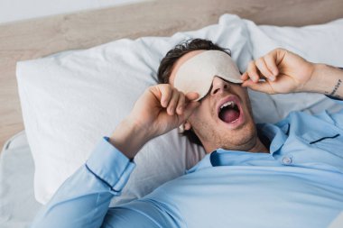 Yatakta esneyen pijama ve uyku maskeli genç adam. 