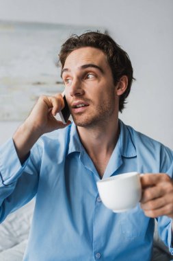 Elinde kahve olan bir adam yatak odasında akıllı telefondan konuşuyor. 