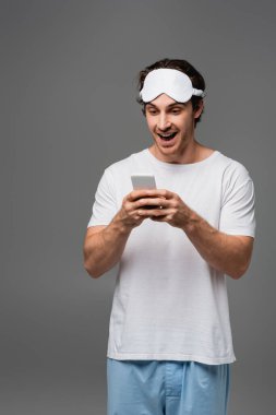 Gri renkte izole edilmiş cep telefonu kullanan uyku maskeli ve pijamalı adam. 