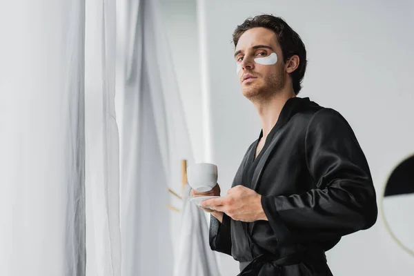 バスルームでカーテンの近くにコーヒーを保持サテンのローブと目のパッチの男の低角度ビュー — ストック写真
