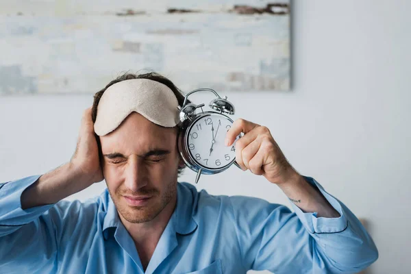 Man in pajama covering ear and holding alarm clock at home