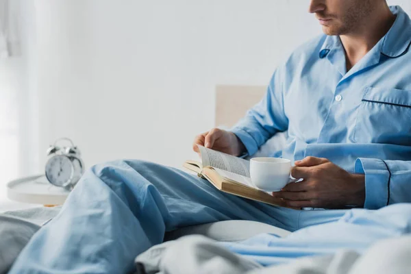 Обрезанный Вид Человека Пижаме Чашкой Книгой Чтения Возле Размытого Будильника — стоковое фото