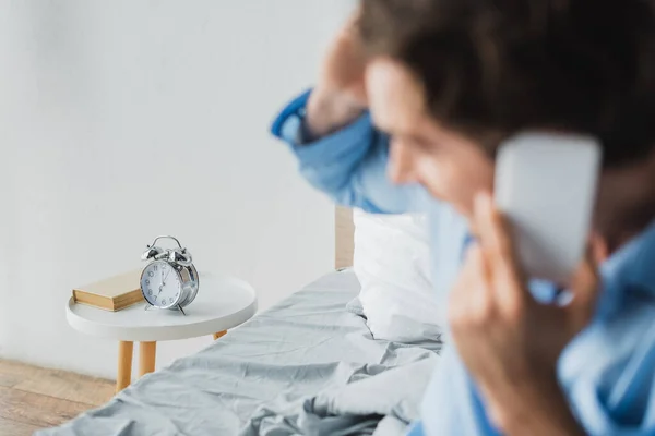 Väckarklocka Och Boka Nattduksbord Nära Suddig Man Pratar Smartphone Sängen — Stockfoto