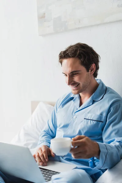 Smiling Freelancer Pajama Using Laptop Holding Blurred Coffee Cup Bed — Stok Foto