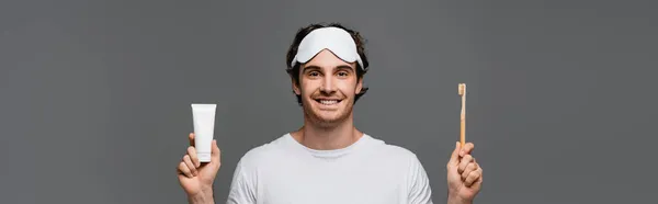 歯ブラシと歯磨き粉を保持しているスリープマスクの笑顔男は灰色で隔離され バナー — ストック写真