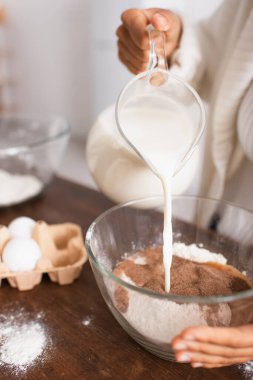 Afro-Amerikalı bir kadının un ve kakaoya süt dökerken çekilmiş görüntüsü. Mutfakta bulanık yumurtalar var. 