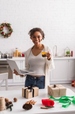 Gülümseyen Afrikalı Amerikalı kadın hediye kutuları ve Noel dekorunun yanında çay ve laptop tutuyor. 