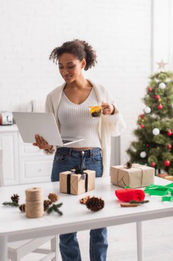 Genç Afrikalı Amerikalı kadın elinde dizüstü bilgisayar ve çayla hediyeler ve evde Noel dekoru tutuyor. 