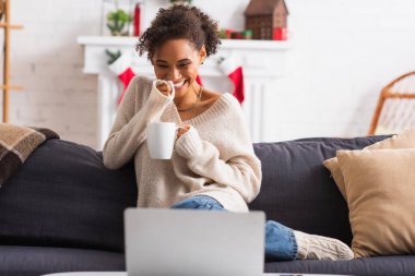 Neşeli Afro-Amerikalı kadın elinde kupayla evde geçirdiği Noel 'de bulanık laptopa bakıyor. 