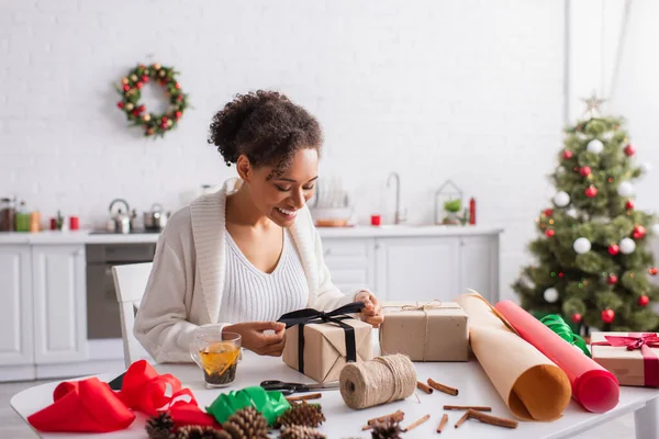 幸せなアフリカ系アメリカ人女性は自宅でお茶や包装紙の近くに贈り物を飾る — ストック写真