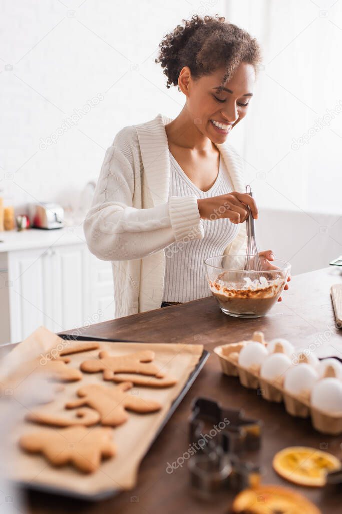 Happy african american woman cooking near blurred eggs and tasty christmas cookies in kitchen 