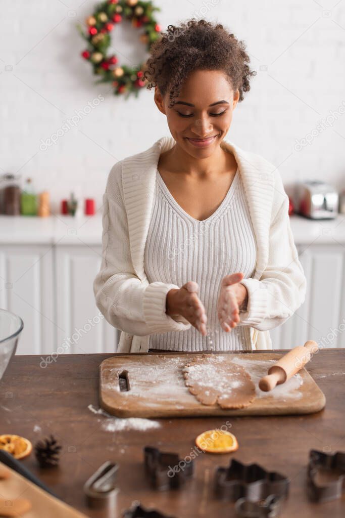 Smiling african american woman pouring flour on dough near rolling pin and cookie cutters in kitchen 