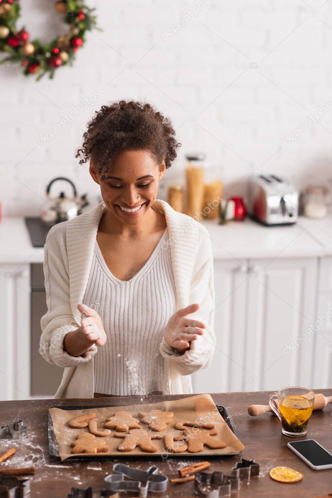 Smiling african american woman pouring flour on christmas cookies near smartphone and tea 