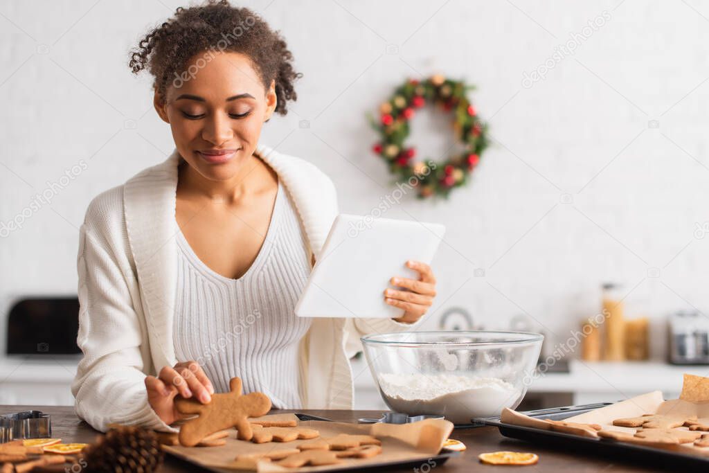 African american woman holding digital tablet near gingerbread and flour in kitchen 