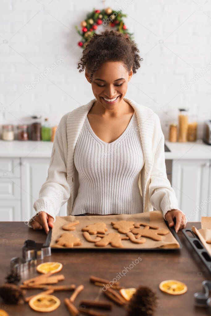Happy african american woman holding tray with christmas gingerbreads near cinnamon sticks 