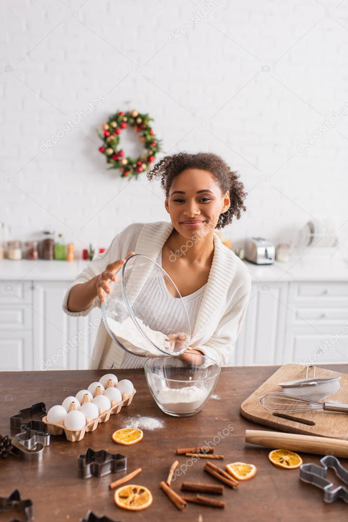 Smiling african american woman pouring flour near ingredients, cinnamon sticks and dry orange in kitchen 
