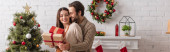 radostná žena se zavřenýma očima drží dárek poblíž manžela a vánoční stromeček, prapor