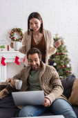 vzrušený pár s čajovými šálky při pohledu na notebook v obývacím pokoji s vánoční výzdobou