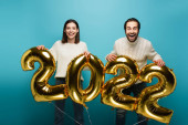 veselý pár dívá na kameru, zatímco drží zlaté balónky ve tvaru 2022 na modré