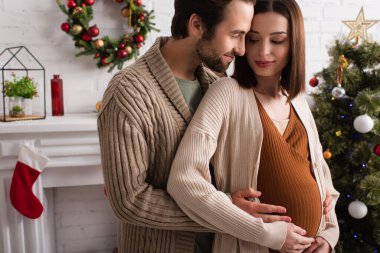 Bulanık Noel ağacının yanında hamile karısını kucaklayan neşeli bir adam.