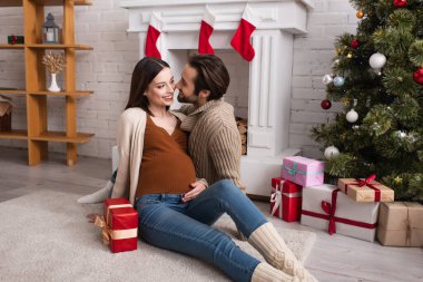 Mutlu hamile kadın ve kocası yerde oturmuş süslü şöminenin yanında ve noel ağacının altında hediyeler veriyorlar.