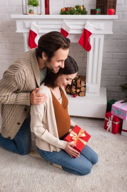 Mutlu bir adam, hamile bir kadının omuzlarına sarılıyor şöminenin yanında oturuyor ve Noel süslemeleri yapıyor.