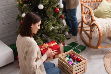 Güler yüzlü bir kadın elinde ahşap kutunun yanında mücevher ve bulanık bir kocayla Noel ağacı süsler.