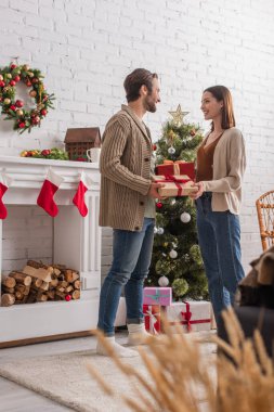 Noel ağacı ve süslü şöminenin yanında hediye kutuları olan mutlu çiftin tam boy görüntüsü.