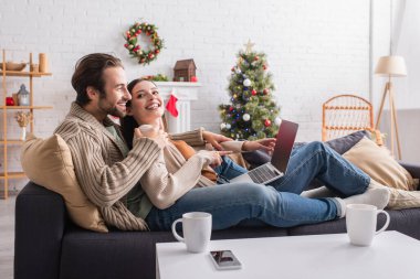 Kocasıyla birlikte oturma odasında Noel süslemeleri ile kanepede dinlenirken dizüstü bilgisayarı gösteren neşeli bir kadın.