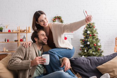 Kocasıyla selfie çeken neşeli kadın oturma odasındaki bulanık Noel ağacının yanında.