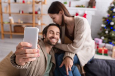 Akıllı telefonun seçici odak noktası mutlu bir adamın eşiyle dekore edilmiş oturma odasında selfie çekmesi.