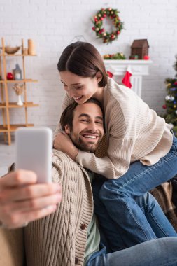 Neşeli bir kadın kocasını kucaklıyor. Süslü oturma odasında bulanık akıllı telefondan selfie çekiyor.