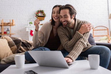 Gülümseyen adam, karısının yanında dizüstü bilgisayar ve kredi kartıyla oturma odasında Noel süslemesi yapıyor.