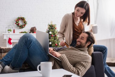 Karısı kredi kartıyla dizüstü bilgisayarda yazan mutlu bir adam oturma odasında Noel süslemeleri ile birlikte.