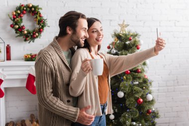 Kocasıyla bulanık noel ağacının yanında selfie çekerken elinde çay tutan neşeli kadın.