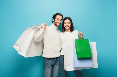 Örgü kazaklı mutlu bir çift alışveriş torbası tutuyor ve mavi kameraya bakıyor.