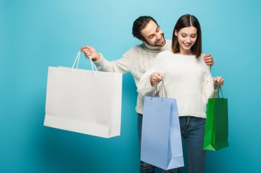 Gülümseyen adam, mutlu bir kadının yanında elinde mavi poşetler tutuyor.